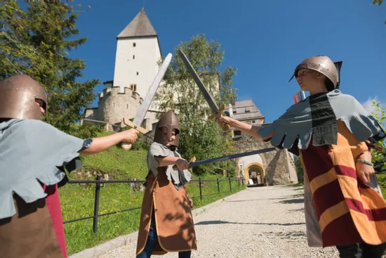Kinder in mittelalterlichen Kostümen spielen Schwertkampf vor einer historischen Burg an einem sonnigen Tag
